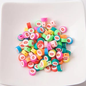 Peut inclure: Un ensemble de perles colorées en pâte polymère, de formes variées, avec un motif de lèvres rouges répété sur fond blanc. Les perles sont de couleurs rose, bleu, vert, orange et jaune, présentées sur une surface blanche.