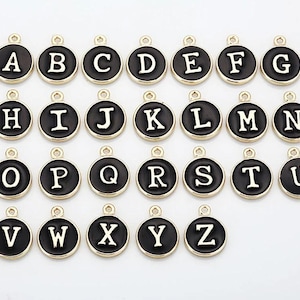 Könnte beinhalten: Eine Sammlung von 26 Buchstabenanhängern. Jeder Anhänger hat einen schwarzen Emaille-Hintergrund mit einem goldfarbenen Rand und einem weißen Buchstaben. Die Anhänger sind rund und haben oben eine kleine Schlaufe zum Befestigen.