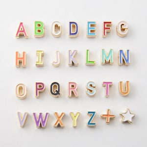 May include: Colorful alphabet letter charms in various colors, including pink, green, blue, and orange. Each letter has a gold-colored border. The letters are arranged in rows, with a plus sign and a star charm at the end.