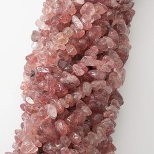 May include: Close-up of a strand of pink strawberry quartz gemstone beads. The beads are small, irregular shapes with a translucent appearance and a mix of pink and clear tones. The background is plain white.