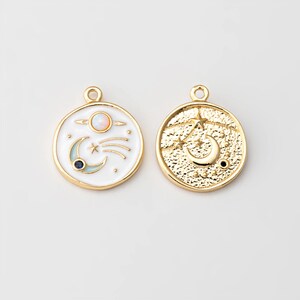 May include: Two gold-toned celestial charms. One charm features a white background with a planet, shooting star, crescent moon, and a blue gem. The other charm has a textured gold surface with a crescent moon and stars.