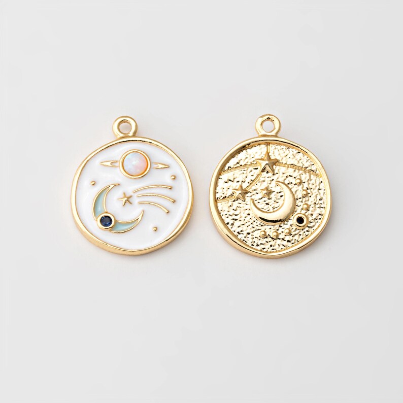 May include: Two gold-toned celestial charms. One charm features a white background with a planet, shooting star, crescent moon, and a blue gem. The other charm has a textured gold surface with a crescent moon and stars.