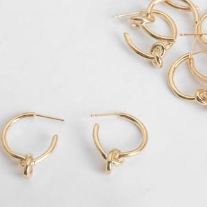 May include: Gold-colored hoop earrings with a knot design. The earrings are on a white background. Several pairs of earrings are visible, with two pairs in the foreground and others scattered in the background. The earrings have a simple, elegant design.