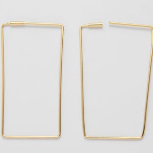 Könnte beinhalten: Zwei goldfarbene, rechteckige Creolen. Die Ohrringe bestehen aus dünnen, geraden Metallstäben, die eine geometrische Form bilden. Ein Ohrring ist vollständig montiert, der andere ist teilweise geöffnet.