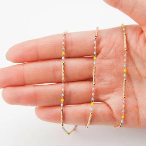 May include: Delicate gold necklaces with colorful beads in pastel shades of pink, yellow, and blue. The necklaces feature vertical gold bars and are held in a hand against a white background. The jewelry is elegant and suitable for everyday wear.