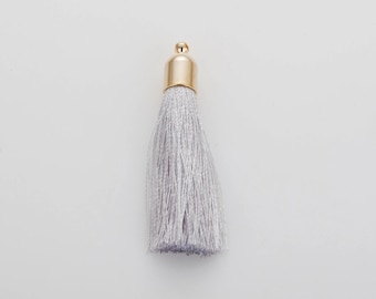 May include: A single, silver-gray tassel with a gold-colored cap. The tassel is made of many strands of thread, and the cap is smooth and shiny. The tassel is a decorative item, likely for jewelry or crafts.