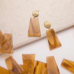 Könnte beinhalten: Ohrringe aus braunem und beigem Harz mit einem goldfarbenen Spiraldesign. Die Ohrringe haben eine rechteckige Form mit einem leichten Trapezdesign. Mehrere lose Ohrringkomponenten sind verstreut.