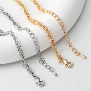 May include: Close-up of three metal chain extenders in gold and silver tones. The chains have different link styles, including textured and oval links. Each extender has a lobster clasp or a teardrop-shaped end for attachment.