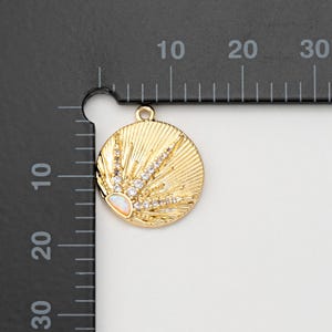 May include: A gold-colored round pendant with a sunburst design. The pendant features a central opal-like stone and is embellished with clear crystals. The pendant is approximately 1 inch in diameter.