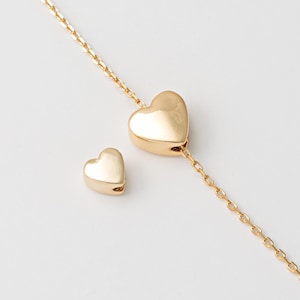 Op de afbeelding: Een goudkleurige ketting met twee hartvormige hangers. Eén hart is bevestigd aan een fijne ketting, de andere is een losse bedel. De sieraden worden gepresenteerd tegen een witte achtergrond.