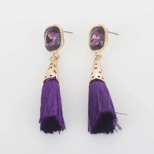 May include: A pair of drop earrings with a gold-coloured setting. Each earring features a square, purple gemstone, a decorative gold-coloured cone, and a long, purple tassel. The earrings are displayed against a white background.