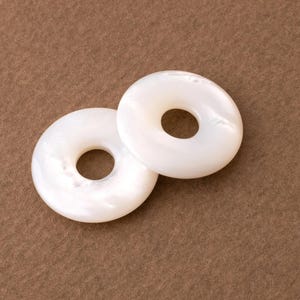 May include: Two white, donut-shaped beads with a pearlescent sheen. The beads have a circular hole in the center and are set against a brown background. These beads could be used for jewelry making or other craft projects.