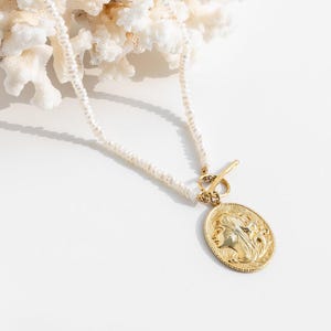 May include: A white pearl necklace with a gold pendant featuring a woman's face. The pendant is attached to the necklace with a gold toggle clasp.