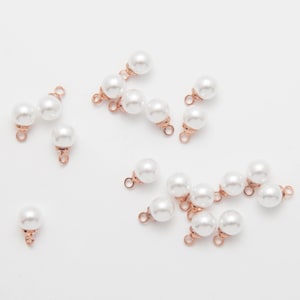 May include: A collection of white faux pearl beads with rose gold-colored metal findings. The beads are round and have a glossy finish. Some beads are connected in pairs or trios, while others are single.