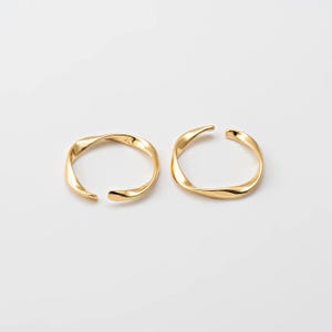 Pode incluir: Dois anéis dourados de extremidade aberta com um design torcido. Os anéis são feitos de um metal brilhante e são colocados sobre um fundo branco. Os anéis têm uma superfície lisa e polida e um estilo moderno e minimalista.