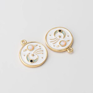 May include: Two gold-toned round charms with a celestial design. Each charm features a white enamel background with a crescent moon, stars, and a planet with an opal-like center. The charms have a loop for attaching to jewelry.
