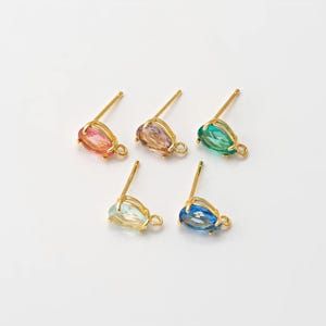 Könnte beinhalten: Fünf Paar Ohrstecker mit tropfenförmigen, farbigen Edelsteinen. Jeder Ohrring hat einen goldfarbenen Stift und eine kleine Schlaufe am Fuß des Edelsteins. Die Edelsteine sind in den Farbtönen Rosa, Lila, Grün, Hellblau und Dunkelblau gehalten.