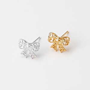 May include: Two bow-shaped stud earrings. One is silver-colored, the other is gold-colored. Both earrings have a delicate, openwork design with heart-shaped cutouts. The earrings are displayed on a white background.