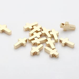 Könnte beinhalten: Eine Sammlung kleiner, goldfarbener Kreuzanhänger. Jeder Anhänger hat ein schlichtes, blockartiges Design mit einem Loch oben zum Auffädeln. Die Kreuze sind auf einer weißen Oberfläche verstreut und erzeugen eine minimalistische Ästhetik.