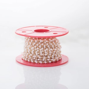 Puede incluir: Un carrete de cadena de perlas con eslabones dorados. La cadena está enrollada alrededor de un carrete de plástico rosa brillante. Las perlas son pequeñas y redondas, creando una apariencia delicada y elegante. Ideal para la fabricación de joyas.