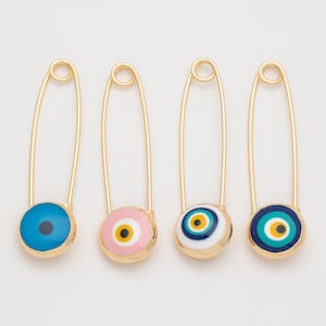 Puede incluir: Cuatro imperdibles dorados con amuletos de ojo turco de colores. Los amuletos son azules, rosas, blancos y azul marino con detalles amarillos y blancos. Los imperdibles están colocados sobre un fondo blanco.