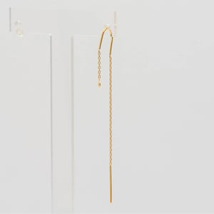 Puede incluir: Un solo pendiente de hilo dorado. El pendiente presenta una parte superior curvada que se transforma en una delicada cadena, que culmina en una barra recta. El pendiente se muestra sobre un fondo blanco.