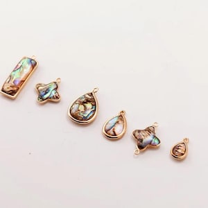 May include: A collection of six gold-toned pendants with iridescent abalone shell inlays. The pendants vary in shape, including a rectangle, a four-leaf clover, teardrops, and a small oval. Each pendant has a loop for attaching to a chain or other jewelry component.