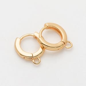 May include: Two gold-colored hoop earrings with a polished finish are displayed against a white background. Each earring has a hinged closure and a small loop for attaching charms or other embellishments. The earrings are simple and elegant.