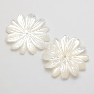Puede incluir: Dos componentes blancos en forma de flor con m&uacute;ltiples p&eacute;talos. Cada componente tiene un agujero central y un brillo nacarado, lo que sugiere n&aacute;car o un material similar. Los p&eacute;talos est&aacute;n sutilmente veteados.