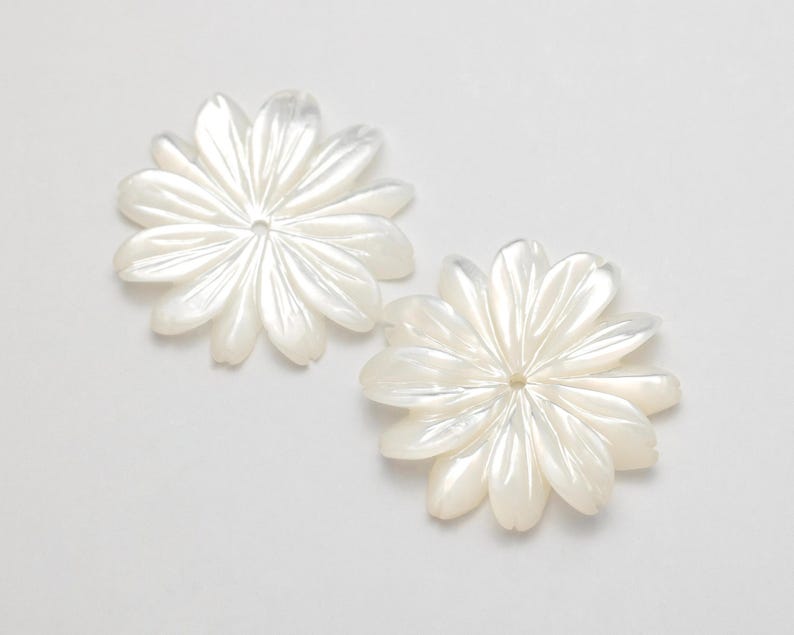Puede incluir: Dos componentes blancos en forma de flor con m&uacute;ltiples p&eacute;talos. Cada componente tiene un agujero central y un brillo nacarado, lo que sugiere n&aacute;car o un material similar. Los p&eacute;talos est&aacute;n sutilmente veteados.