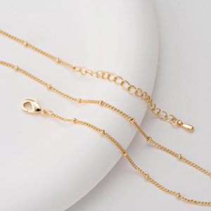 Puede incluir: Un delicado collar dorado con un diseño de cadena de bolitas. El collar presenta un cierre de mosquetón y una cadena ajustable. La joya se exhibe sobre una superficie blanca.