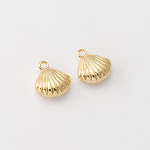 May include: Two gold-colored seashell charms with a loop at the top for jewelry making. The charms have a ridged texture, resembling a real seashell. The background is plain white.