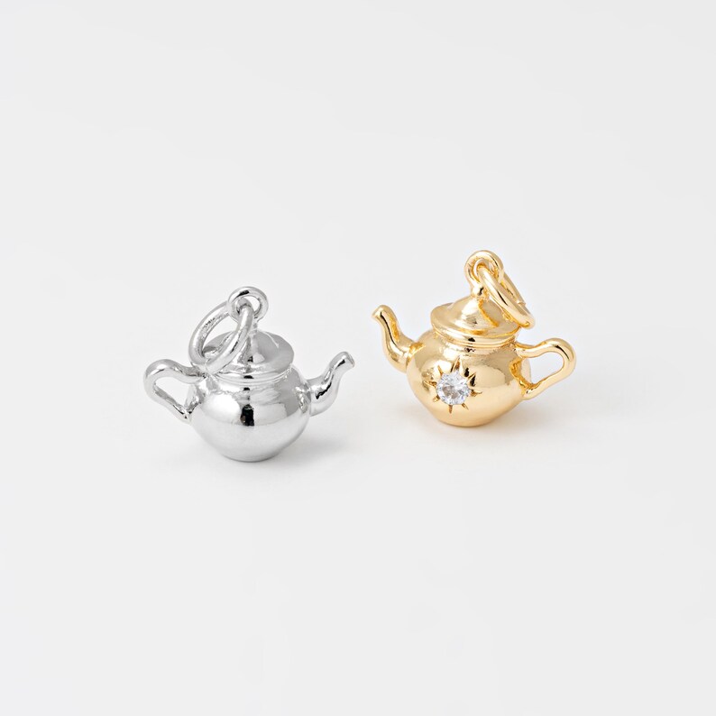 May include: Two small teapot charms, one silver and one gold. The silver teapot has a shiny, reflective surface. The gold teapot features a small, clear gemstone on the front. Both charms have a loop at the top for attaching to a necklace or bracelet.