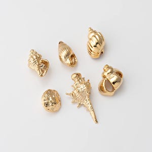 May include: Six gold-colored seashell charms in various shapes and sizes are arranged on a white surface. The charms have detailed textures and are likely made of metal. These decorative charms are suitable for jewelry making or craft projects.