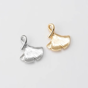 May include: Two metal pendants in the shape of ginkgo leaves. One pendant is silver, and the other is gold. The pendants have a detailed, textured surface and a loop at the top for hanging. The background is plain white.