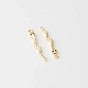 May include: Two gold-coloured snake charms with green eyes. The charms have a wavy, serpentine design and a small loop at the top for attaching to jewellery. The background is plain white.
