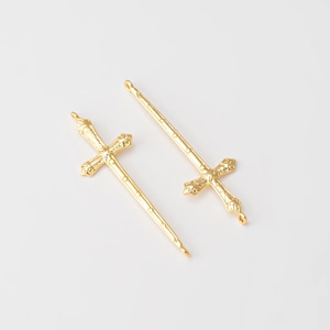 Puede incluir: Dos colgantes dorados con forma de espada y empuñaduras en forma de cruz. Los colgantes están detallados con diseños ornamentados y tienen un lazo en la parte superior para colgar. El fondo es blanco liso.