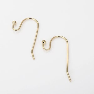 May include: Two gold-colored earring hooks. Each hook has a curved design with a small ball at the top and a straight, pointed bottom. The hooks are displayed against a plain white background.