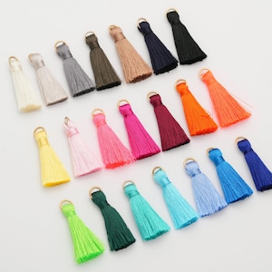 May include: A collection of colorful tassels in various shades, including white, gray, pink, orange, and blue. Each tassel has a gold-colored metal loop at the top. These decorative tassels are ideal for jewelry making or craft projects.
