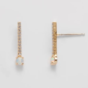 May include: A pair of gold-colored stud earrings. Each earring features a vertical bar encrusted with small, clear stones and a round, opalescent stone at the bottom. The earrings are set against a plain, light background.