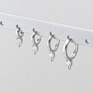 May include: Four silver hoop earrings with a dangling clear gemstone. The earrings are displayed on a silver bar with small holes. The gemstones are teardrop-shaped and catch the light.