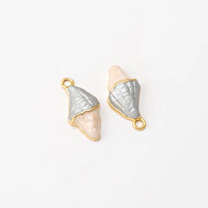May include: Two seashell-shaped charms. Each charm has a light pink base, a silver-colored top, and gold-colored trim. The charms have a small gold loop at the top for attaching to jewelry. The background is white.