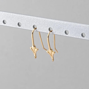 Puede incluir: Un par de pendientes colgantes dorados. Cada pendiente presenta un diseño floral decorativo y estilizado en la parte inferior y un gancho curvo para orejas perforadas. Los pendientes se muestran sobre un fondo gris claro.