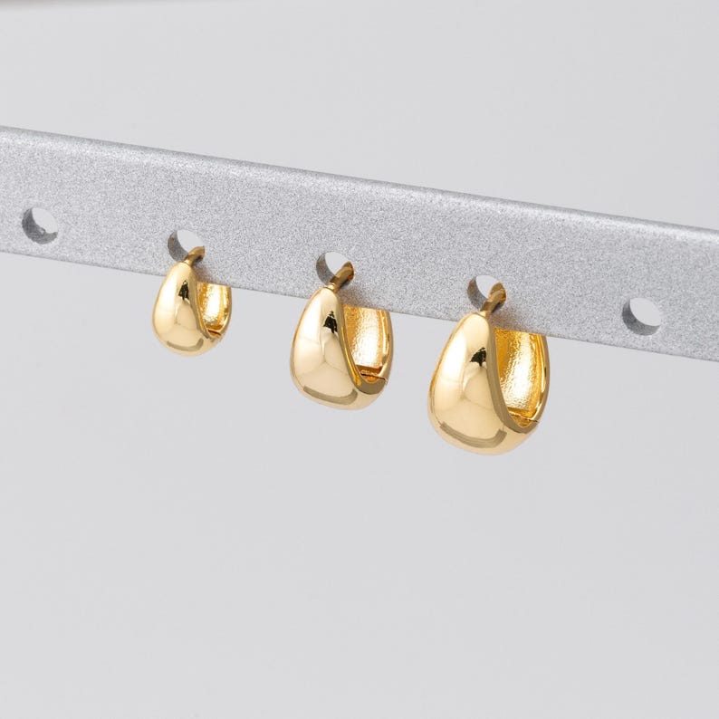 May include: Three gold-coloured hoop earrings of varying sizes are displayed on a silver bar. The earrings have a smooth, polished finish and a rounded, chunky design. The earrings are arranged in a row, showcasing their different sizes.