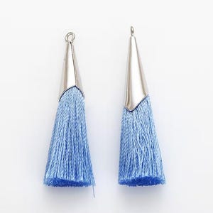 May include: Two light blue tassel pendants with silver-colored metal caps. The tassels are made of fine threads, and the metal caps have a loop at the top for hanging. The pendants are set against a white background.