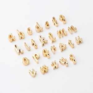 May include: Gold-colored metal alphabet charms in a cursive font. The charms are arranged on a white surface. The letters include a, b, c, d, e, f, g, h, i, j, k, l, m, n, o, p, q, r, s, t, u, v, w, x, y, and z.