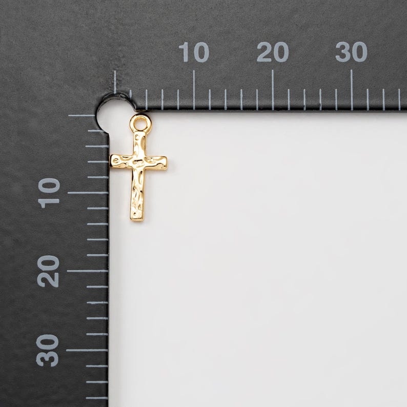 Puede incluir: Un peque&ntilde;o colgante de cruz de color dorado con una superficie texturizada. La cruz tiene un lazo en la parte superior para sujetarlo a una cadena u otra joyer&iacute;a. El colgante se muestra sobre un fondo de regla en blanco y negro.