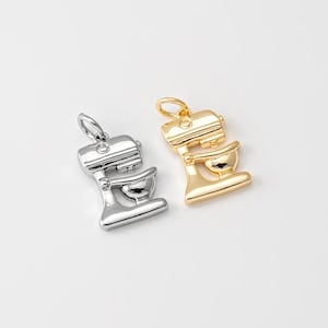May include: Two metal mixer charms, one silver and one gold, are displayed against a white background. The charms are detailed miniature replicas of stand mixers, featuring a bowl and a top section with a loop for hanging. The gold charm has a shiny finish.