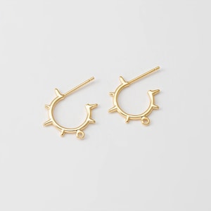 Op de afbeelding: Een paar goudkleurige oorringen. Elke oorring heeft een cirkelvormig ontwerp met kleine, gelijkmatig verdeelde spikes rond de buitenrand. De oorbellen hebben een pin voor piercing en een kleine lus aan de onderkant.