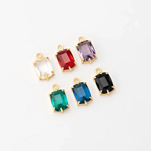 May include: Seven rectangular gemstone charms in various colors: clear, red, purple, green, blue, and black. Each gem is set in a gold-colored metal frame with a small loop at the top for attaching to jewelry. The charms are arranged on a white surface.